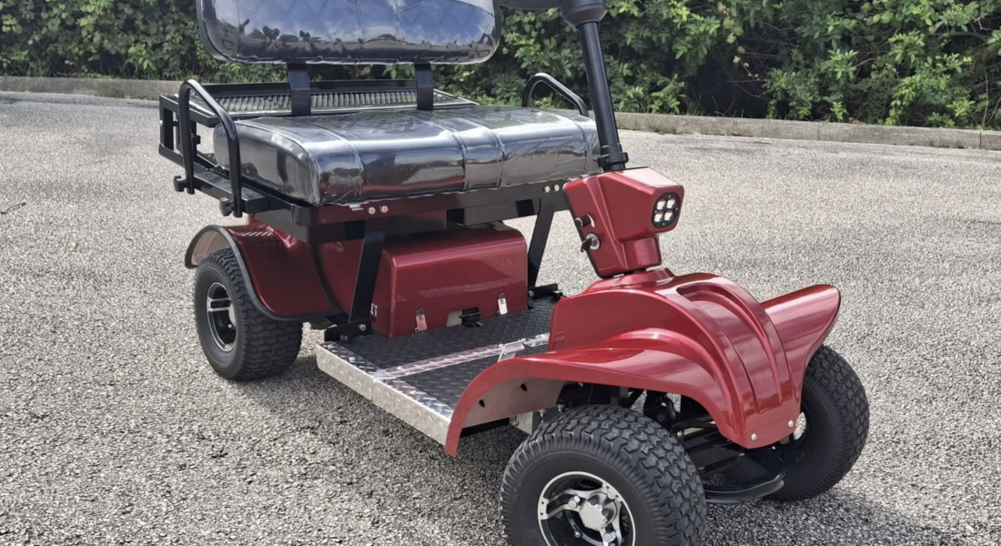 Cricket-mini-cart-model-SR1 Red mobility scooter parked on a paved lot with a black cushioned seat and large wheels, hedge in the background.